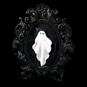 Peut inclure: Un fantôme blanc avec deux yeux noirs est centré dans un cadre ovale noir orné. Une toile d'araignée est visible derrière le fantôme. Le cadre a un design baroque détaillé. Le fond est noir uni, créant un thème d'Halloween.