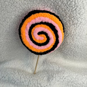 May include: A colorful, handmade lollipop with a spiral pattern in black, orange, and pink yarn. The lollipop is attached to a wooden stick.