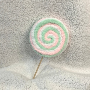 May include: A pink and green swirl lollipop on a wooden stick. The lollipop is on a white fluffy surface.