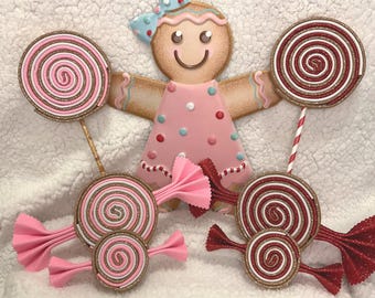 May include: A gingerbread girl with a pink dress and a blue bow, surrounded by decorative lollipops and candy. The lollipops have pink and white swirls with gold glitter accents. The candy has red and pink bows.