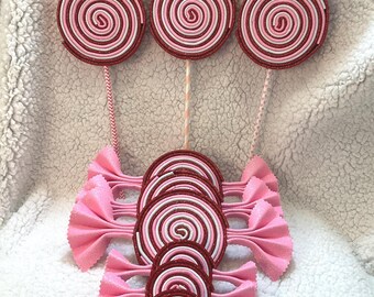 May include: A set of pink and red swirl lollipop decorations. The lollipops are made of felt and have a white swirl in the center. There are also pink felt bows with a red swirl lollipop in the center.