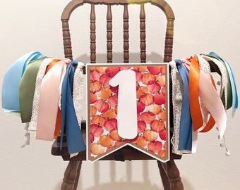 May include: A pennant banner featuring a watercolor pumpkin pattern in shades of orange, red, and yellow with a large white number one in the center. The banner is decorated with multiple hanging ribbons in blue, orange, pink, and white lace.