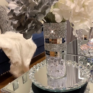 May include: A silver glitter vase with a mirrored band and clear glass beads sits on a mirrored tray with a crystal trim. The vase is filled with gray and white silk flowers.