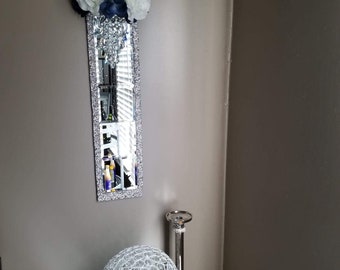 Può includere: Uno specchio con cornice argentata con un design a lampadario di cristallo e un'arrangiamento floreale bianco e blu navy in cima. Lo specchio è appeso a una parete marrone chiaro. Una lampada da tavolo argentata con un paralume a globo di cristallo è su un tavolo argentato di fronte allo specchio.