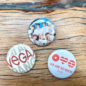 May include: Three circular pin-back buttons. One features a group of seven people in white shirts against a blue sky. Another is white with red text reading VEGAS. The third is white with red symbols and the text WE ARE SO BACK 2026.