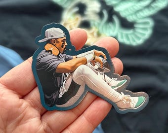 May include: A sticker featuring a baseball player in a seated pose, wearing a gray shirt, white pants, and a white cap. The player has a tan neckband and white wristbands. The sticker has a blue border and the shoes have teal accents.