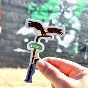 May include: A white sticker with a brown and white bald eagle perched on a streetlight. The streetlight has a green sign that reads "BROAD ST".
