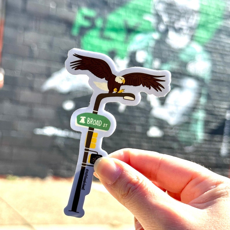 May include: A white sticker with a brown and white bald eagle perched on a streetlight. The streetlight has a green sign that reads "BROAD ST".