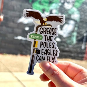 May include: A white sticker with a cartoon eagle perched on a street sign that reads "Grease the Poles, Eagles Can Fly." The street sign also has a small bell and reads "Broad St."