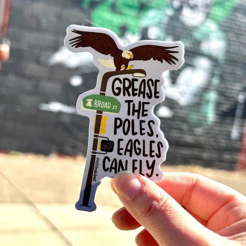 May include: A white sticker with a cartoon eagle perched on a street sign that reads "Grease the Poles, Eagles Can Fly." The street sign also has a small bell and reads "Broad St."