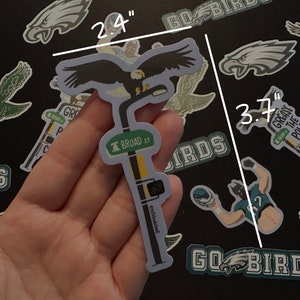May include: A sticker of an eagle perched on a street sign that reads "BROAD ST". The sticker is 2.4 inches tall and 3.7 inches wide. The eagle is black and white, and the street sign is green with white lettering.