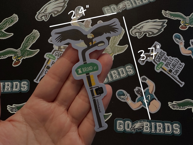May include: A sticker of an eagle perched on a street sign that reads "BROAD ST". The sticker is 2.4 inches tall and 3.7 inches wide. The eagle is black and white, and the street sign is green with white lettering.