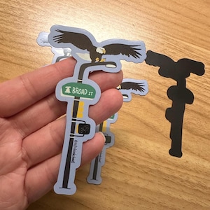 May include: A set of three blue and white stickers featuring a streetlight with a bald eagle perched on top. The streetlight has a green sign that reads "BROAD ST" and a traffic light. The stickers are designed with a vintage aesthetic and are perfect for adding a touch of Americana to any surface.