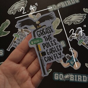May include: A sticker featuring a cartoon eagle perched on a street sign that reads "Grease the Poles. Eagles Can Fly." The sign is yellow with black lettering and has a smaller sign that reads "Broad St." The sticker is 2.4 inches wide and 3.7 inches tall.