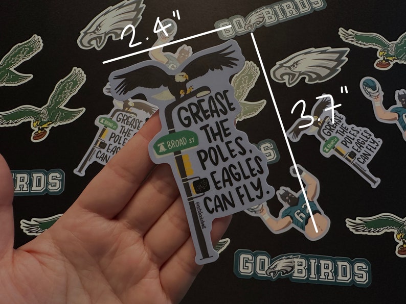 May include: A sticker featuring a cartoon eagle perched on a street sign that reads "Grease the Poles. Eagles Can Fly." The sign is yellow with black lettering and has a smaller sign that reads "Broad St." The sticker is 2.4 inches wide and 3.7 inches tall.