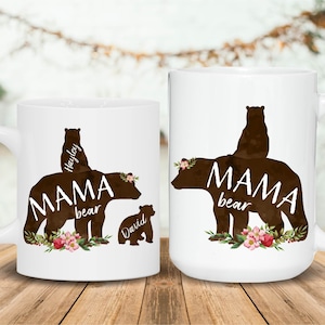 May include: Two white ceramic mugs with brown bear silhouettes. The larger mug has the text "Mama Bear" with a bear cub on top of the mama bear. The smaller mug has the text "Mama Bear" with two bear cubs on top of the mama bear. Both mugs have floral designs.