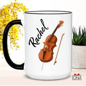 May include: White ceramic mug with a black handle and black rim. The mug has a cello and bow illustration and the name "Rachel" written in black cursive.