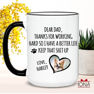 May include: White ceramic coffee mug with a black handle and rim. The mug features a black paw print, a heart with a photo of a corgi, and the text "Dear Dad, Thanks for working hard so I have a better life. Keep that shit up. Love, Harley."