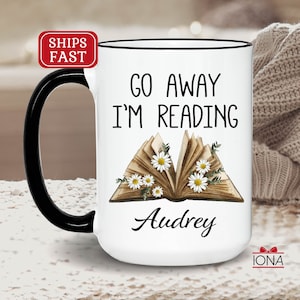 Op de afbeelding: Witte keramische koffiemok met een zwarte handgreep. De mok heeft een zwarte rand en bevat de tekst "Go Away I'm Reading" met een vintage boekillustratie en madeliefjes. De naam "Audrey" staat onder het boek gedrukt.