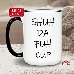 K&ouml;nnte beinhalten: Wei&szlig;e Keramik-Kaffeetasse mit schwarzem Henkel und schwarzem Rand. Die Tasse tr&auml;gt den Schriftzug "SHUH DA FUH CUP" in Schwarz.