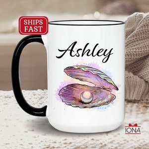 May include: White ceramic mug with a black handle. The mug has a watercolor design of a seashell with a pearl inside. The name "Ashley" is written in black cursive on the mug.