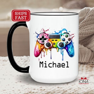 May include: White ceramic mug with a black handle. The mug features a colorful video game controller graphic with dripping paint and the name "Michael" printed below it.