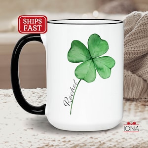May include: White ceramic mug with a black handle. The mug features a green four-leaf clover with a watercolor effect and the name "Rachel" written in cursive.