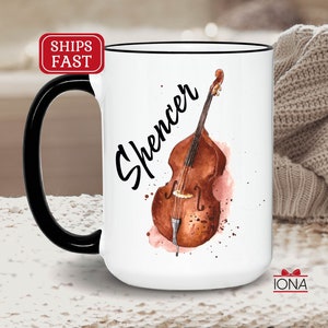 May include: White ceramic mug with a black handle. The mug features a watercolor illustration of a double bass with the name "Spencer" written in black cursive script.