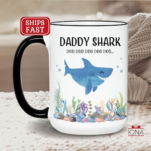 May include: White ceramic coffee mug with a black handle. The mug has a blue shark illustration with the text "Daddy Shark Doo Doo Doo Doo Doo Doo..." printed on it.