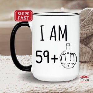 May include: White ceramic coffee mug with a black handle and rim. The mug has black text that reads "I AM 59+ " with a black and white illustration of a middle finger.