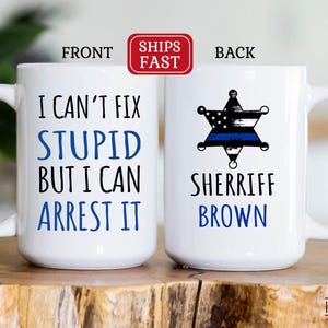 May include: White ceramic mug with the text "I CAN'T FIX STUPID BUT I CAN ARREST IT" on one side and a sheriff's badge with the name "SHERIFF BROWN" on the other. The mug has a large handle.