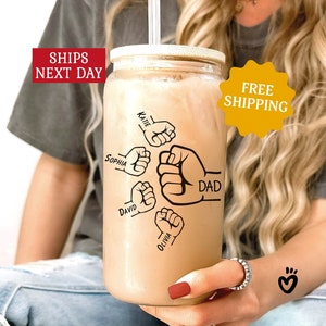May include: A clear glass tumbler with a lid and a straw. The tumbler has a brown liquid inside and black and white illustrations of fists with names written on them. The names are "Katie", "Sophia", "David", "Olivia", and "Dad".
