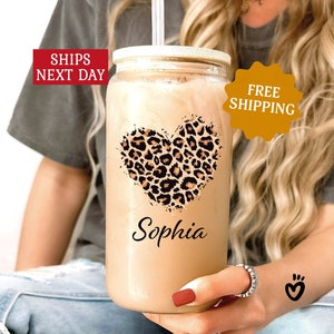 May include: A clear glass tumbler with a lid and a straw. The tumbler has a leopard print heart design and the name "Sophia" printed on it. The tumbler is filled with a light brown liquid.