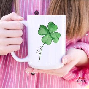 May include: White ceramic mug with a green four-leaf clover design and the name "Tina" written in cursive.