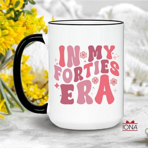 May include: White ceramic mug with a black handle and rim. The mug features the text "IN MY FORTIES ERA" in shades of pink and red, with flower and star accents. The mug is a standard size for coffee or tea.