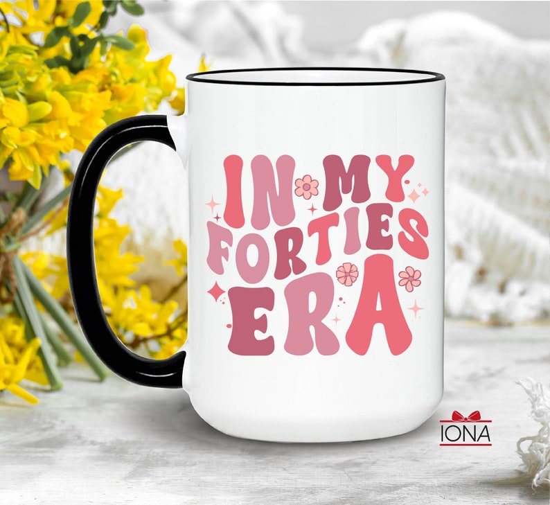 May include: White ceramic mug with a black handle and rim. The mug features the text "IN MY FORTIES ERA" in shades of pink and red, with flower and star accents. The mug is a standard size for coffee or tea.