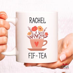 May include: White ceramic mug with a pink teacup and flowers design. The mug says "Rachel Fif-Tea" in black text.