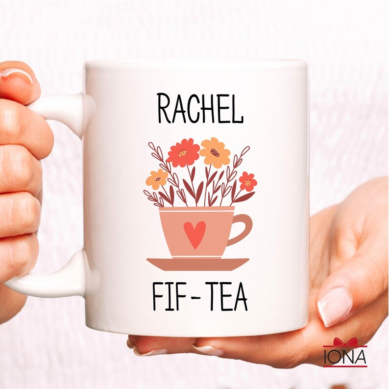 May include: White ceramic mug with a pink teacup and flowers design. The mug says "Rachel Fif-Tea" in black text.
