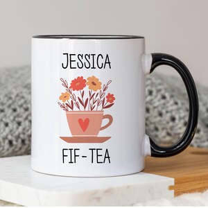 May include: White ceramic mug with a black rim and handle. The mug has a floral design with a teacup and the text "Jessica FIF-TEA".