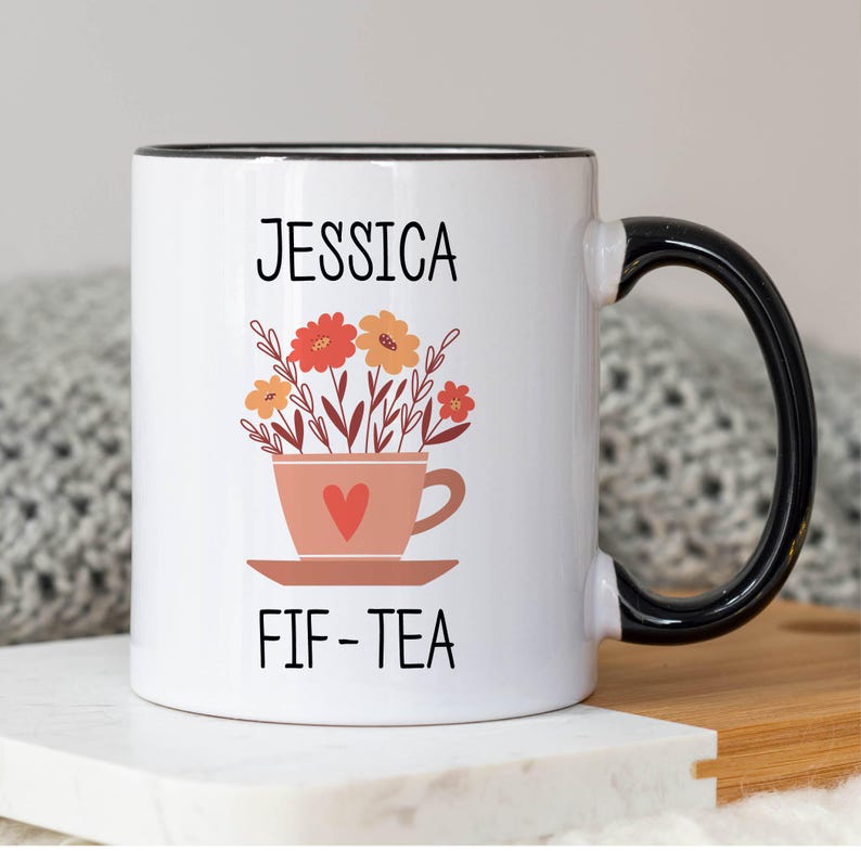 May include: White ceramic mug with a black rim and handle. The mug has a floral design with a teacup and the text "Jessica FIF-TEA".