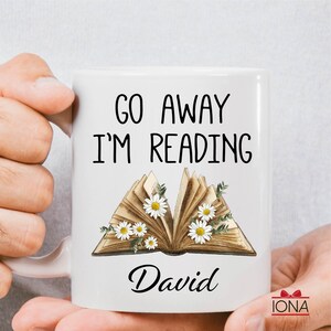 Op de afbeelding: Witte keramische mok met de tekst "Go Away I'm Reading" en een bloemige illustratie van een open boek. De naam "David" staat onder het boek.