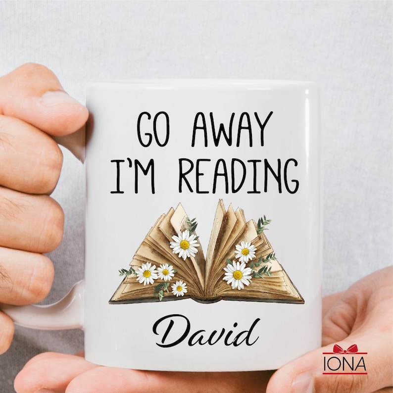 Op de afbeelding: Witte keramische mok met de tekst "Go Away I'm Reading" en een bloemige illustratie van een open boek. De naam "David" staat onder het boek.