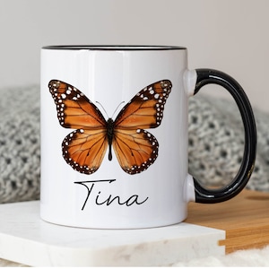 May include: White ceramic mug with a black rim and handle. The mug features a colorful butterfly illustration and the name "Tina" written in cursive.