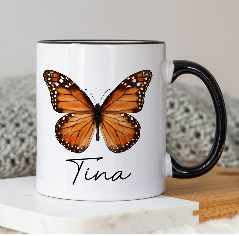May include: White ceramic mug with a black rim and handle. The mug features a colorful butterfly illustration and the name "Tina" written in cursive.
