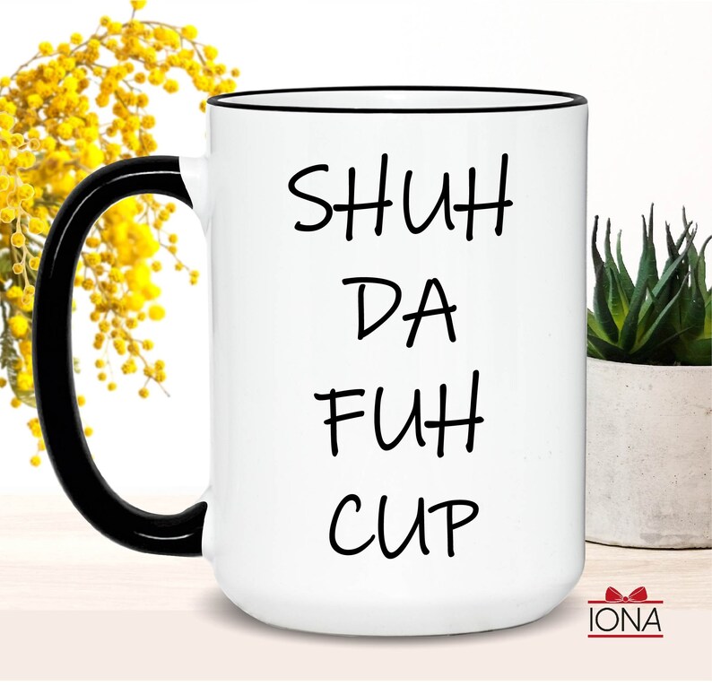 K&ouml;nnte beinhalten: Wei&szlig;e Keramiktasse mit schwarzem Rand und Henkel. Die Tasse hat einen schwarzen Schriftzug mit der Aufschrift "SHUH DA FUH CUP".