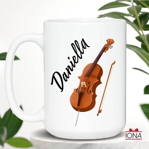 May include: White ceramic mug with a black cursive name "Daniella" and a brown cello with a bow and a bow stick.