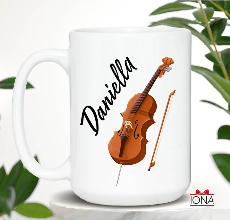 May include: White ceramic mug with a black cursive name "Daniella" and a brown cello with a bow and a bow stick.
