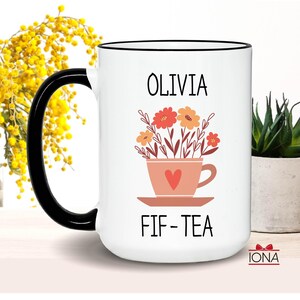 May include: White ceramic mug with a black rim and handle. The mug has a floral design with a teacup and the text "Olivia Fif-Tea".