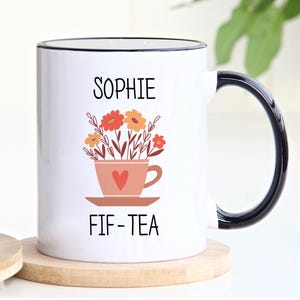 May include: White ceramic mug with a black rim and handle. The mug has a floral design with a teacup and saucer. The text "Sophie FIF-TEA" is printed on the mug.