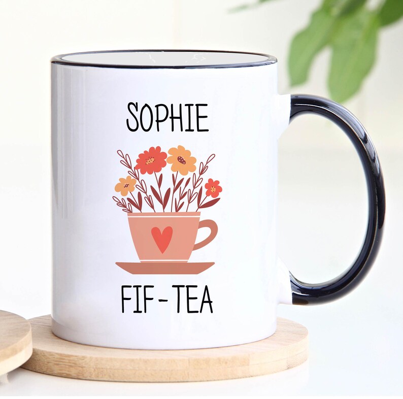 May include: White ceramic mug with a black rim and handle. The mug has a floral design with a teacup and saucer. The text "Sophie FIF-TEA" is printed on the mug.